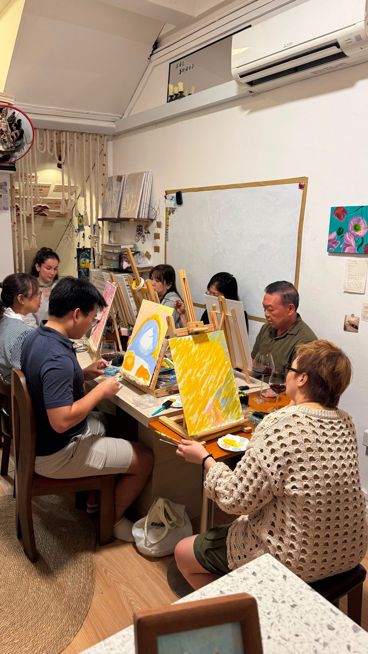 An evening session: people painting at small easels around a long table, with wine glasses between them