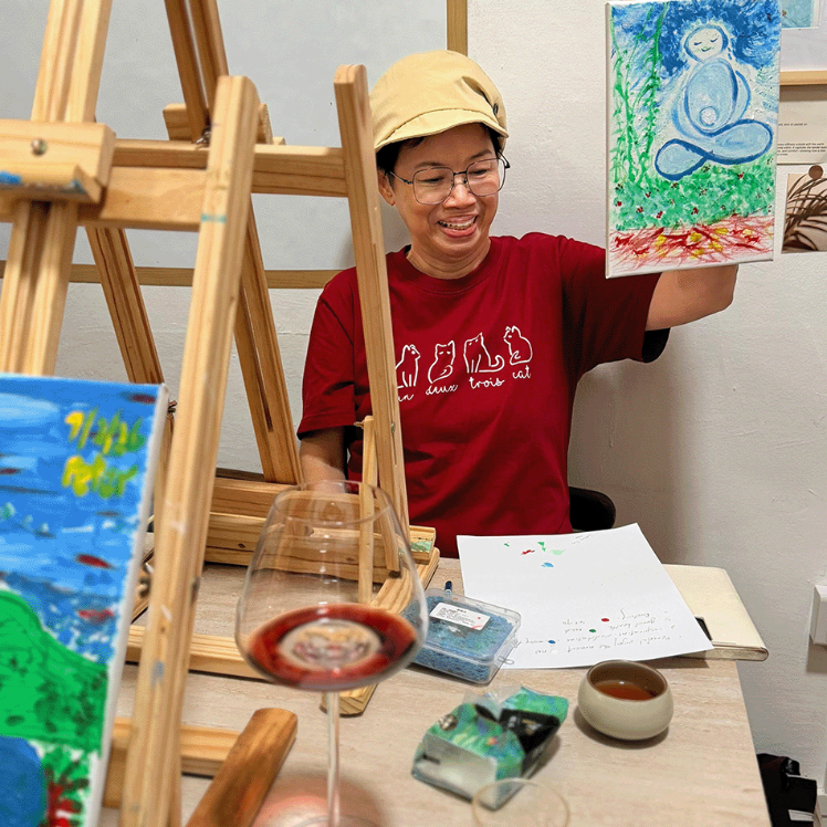 A smiling man wearing a red t-shirt and a beige cap sits behind a wooden easel in an art studio, holding up a colorful painting of a meditating figure. The workspace is cluttered with art supplies, including another easel, a glass of red wine, paint tubes, brushes, and paint palettes.