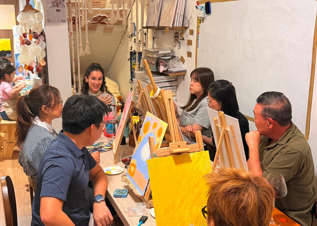 A group of people painting together in a cozy, decorated art studio with warm lighting, easels, and shelves in the background.