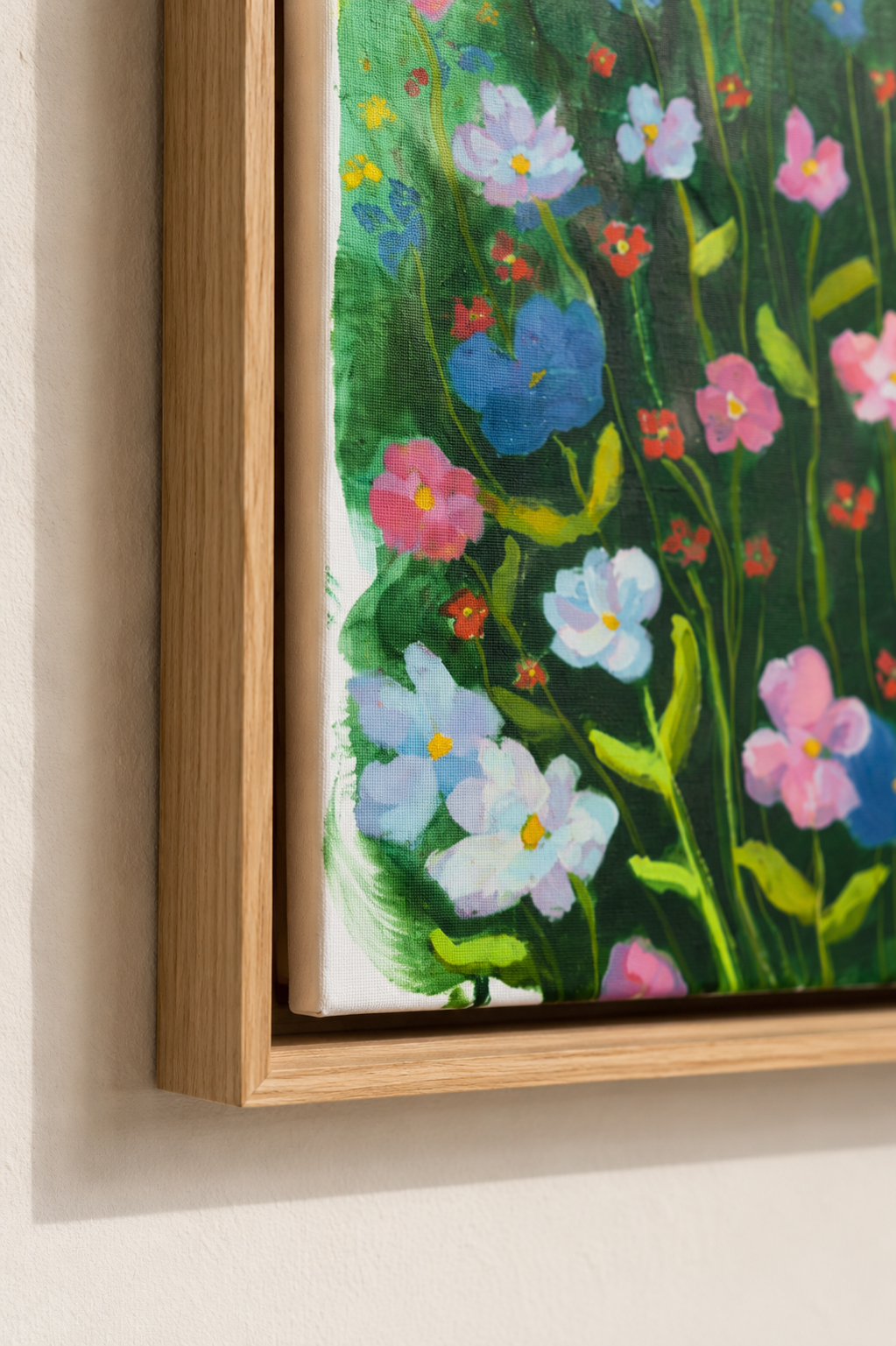 A close-up of a framed floral painting hanging on a light beige wall. The artwork depicts colorful flowers in pink, blue, white, and red with green stems and leaves, enclosed in a light natural wood frame.
