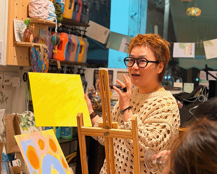 A woman with short auburn hair and glasses stands in a colorful art studio, gesturing toward a yellow canvas on an easel. Other people and colorful hanging lamps are visible in the background and foreground.