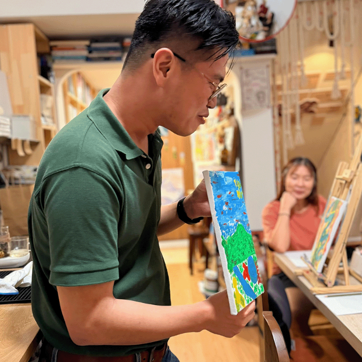 A man in a green polo shirt and jeans holds up a colorful painted canvas showing a blue sky and green landscape in a warm, creative studio environment with another person visible in the background.